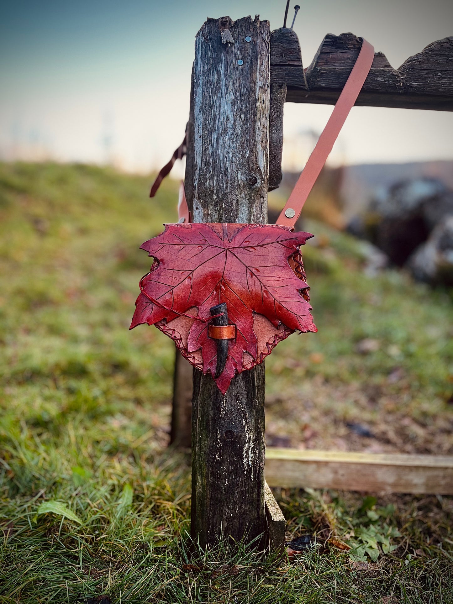 Ahornblatt Ledertasche mit roter Blattklappe und braunem Schulterriemen an einem Holzpfosten.