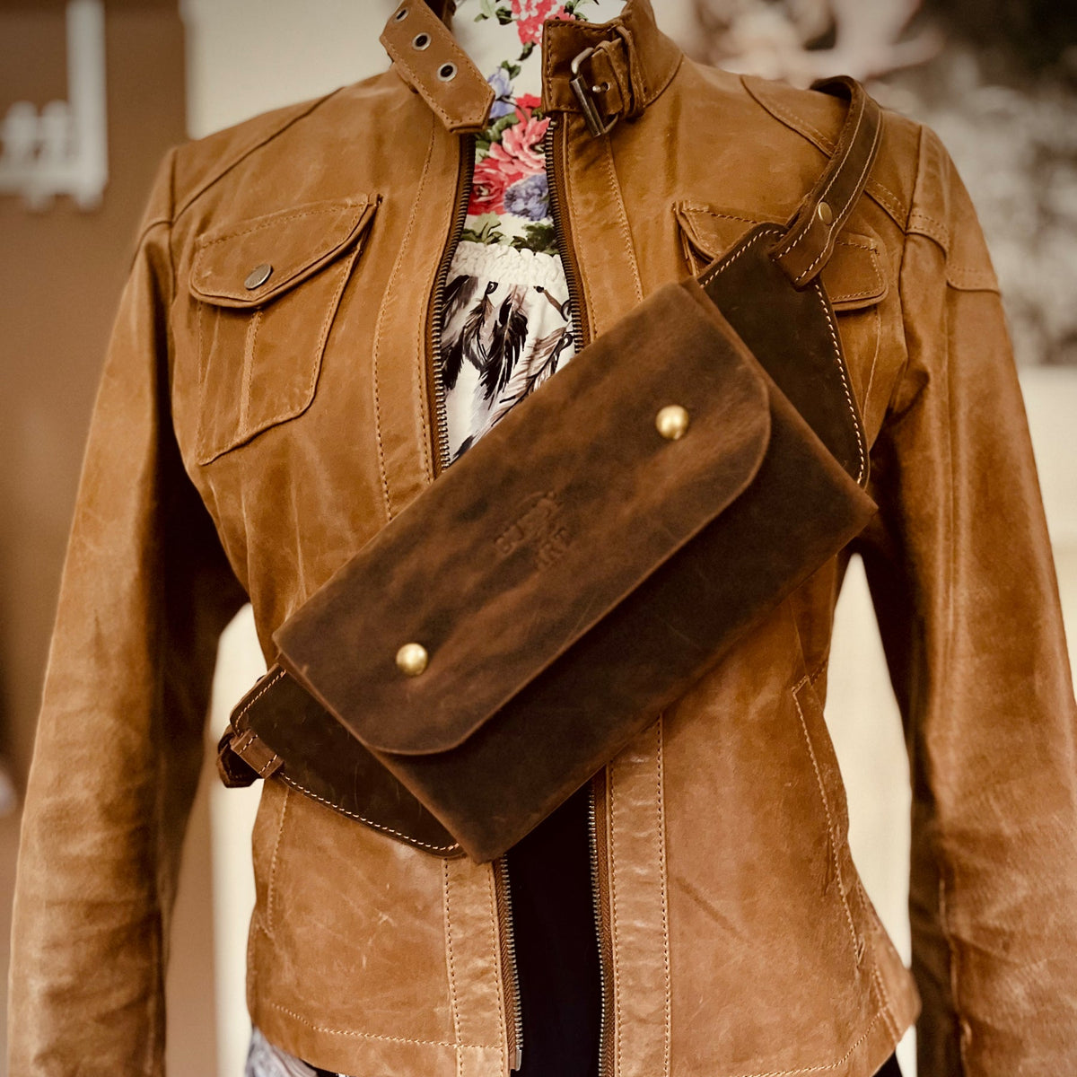 Braune Crossbody Tasche aus Leder mit normaler Klappe, diagonal über einer Lederjacke an einer Schneiderpuppe getragen.