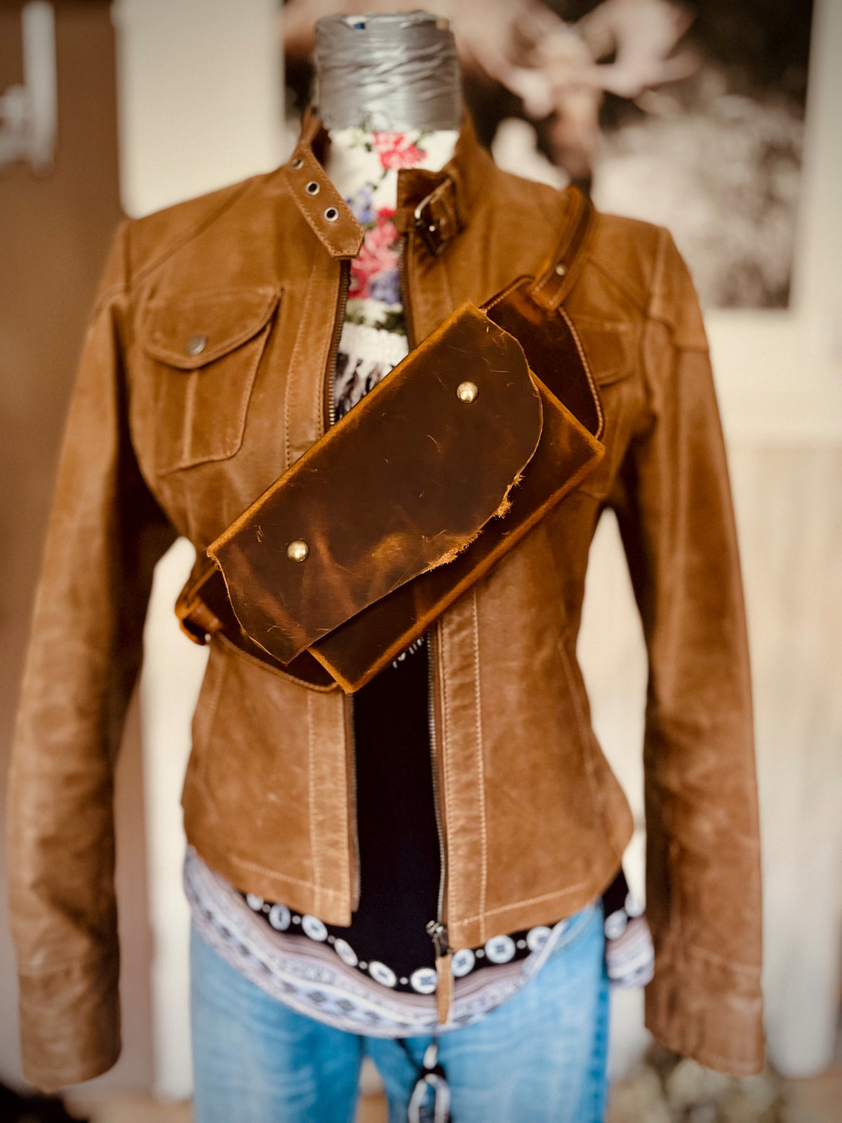 Braune Crossbody Tasche aus Leder mit wilder Klappe, diagonal über einer Lederjacke an einer Schneiderpuppe getragen.