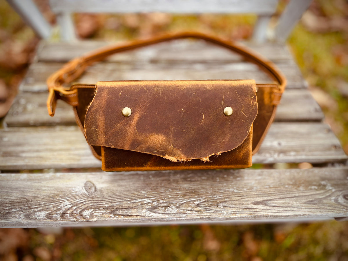 Braune Leder-Bauchtasche mit wilder Klappe und unregelmäßiger Naturkante, frontal auf einer Holzbank.