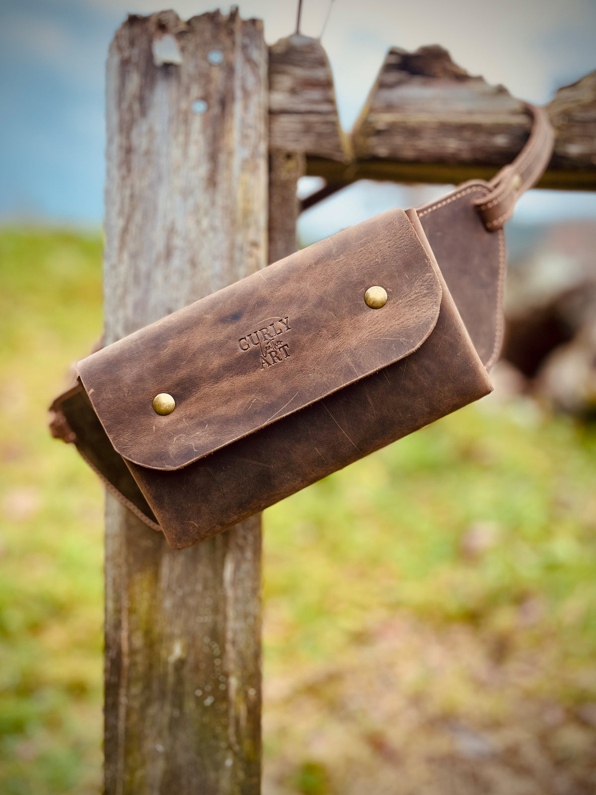 Braune Ledertasche mit normaler Klappe, eingeprägtem Logo und Ledergurt an einem rustikalen Holzpfosten.