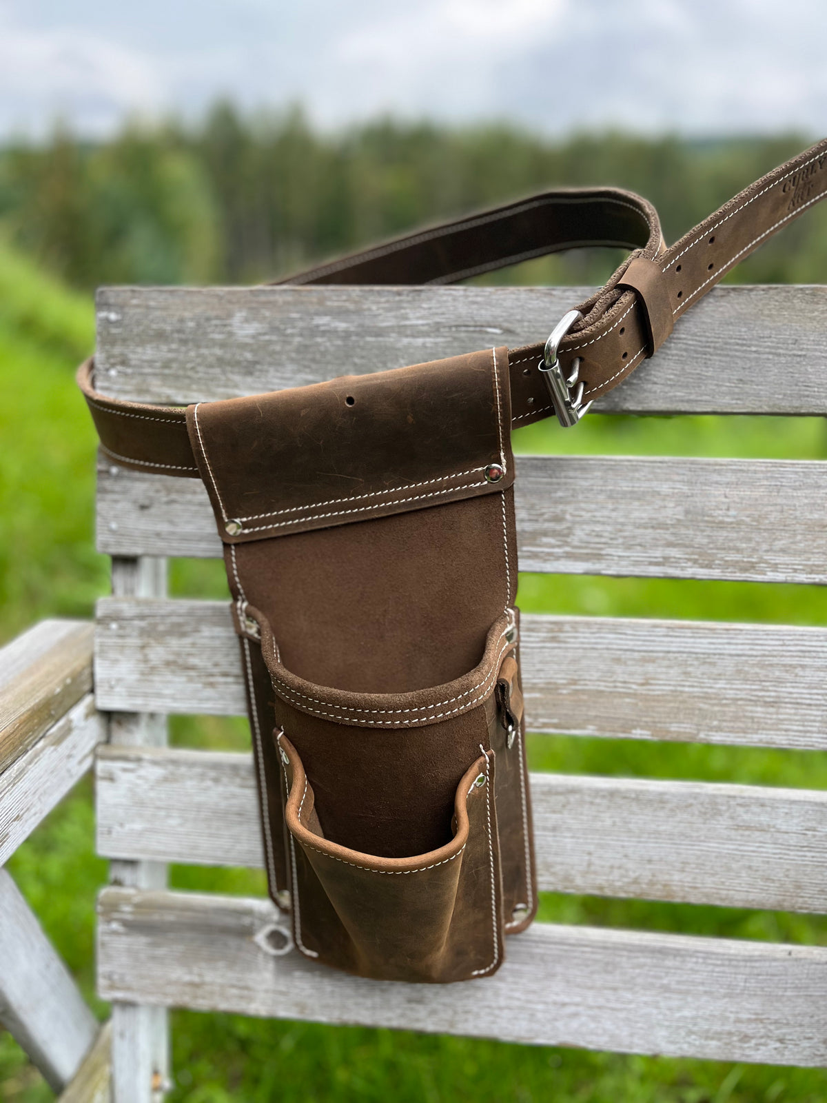 Braune Schraubentasche aus Leder mit Hauptfach und Fronttasche, an einem Ledergürtel auf einer Holzbank