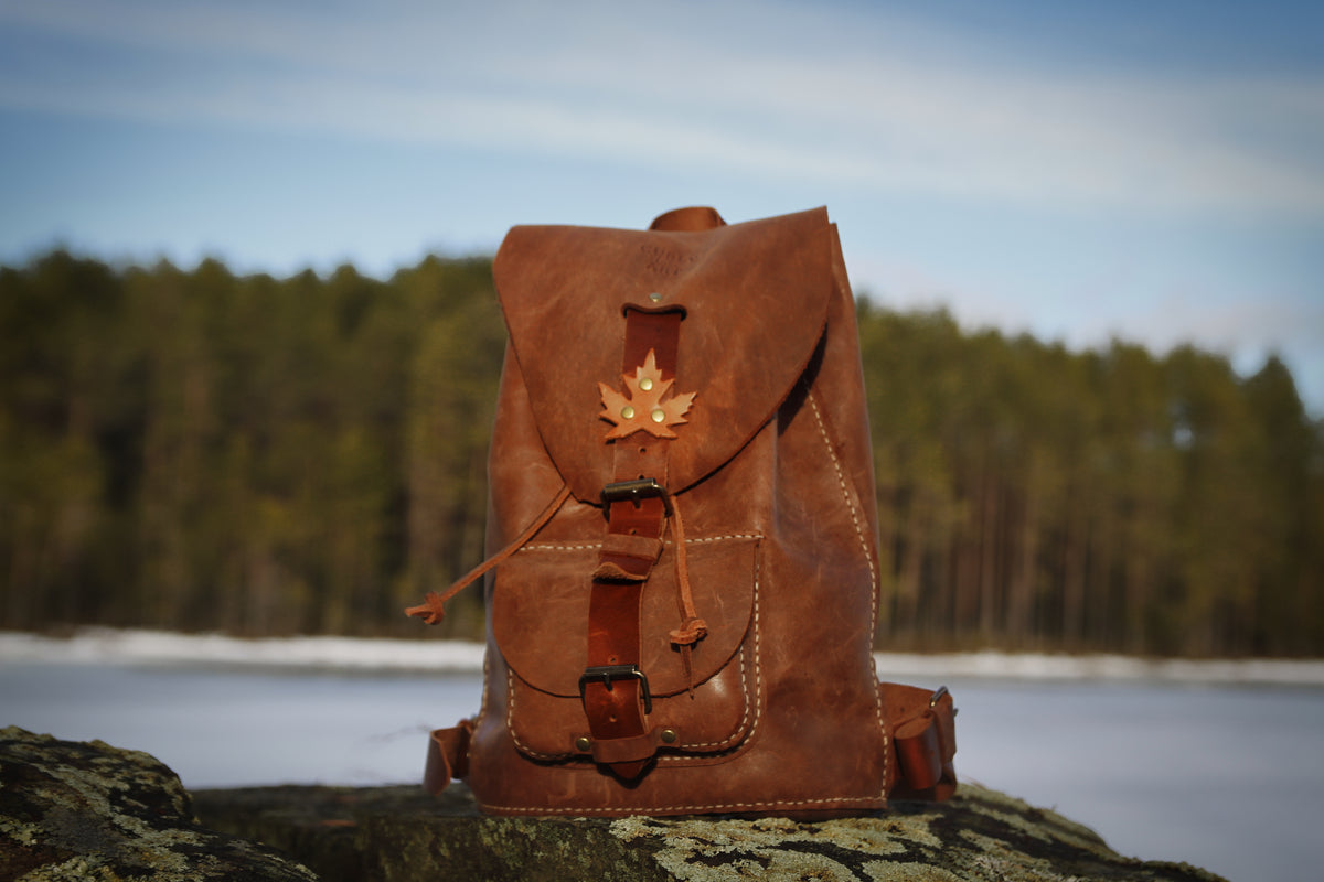 Brauner Lederrucksack mit Ahornblatt und Fronttasche auf einem Felsen vor einem See.