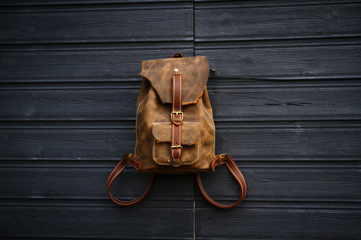 Brauner Lederrucksack mit natürlicher Lederkante und ausgebreiteten Schulterriemen vor einer dunklen Holzwand
