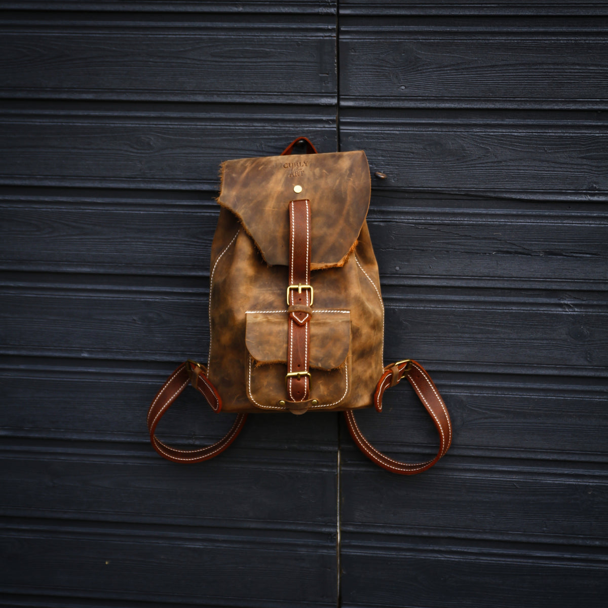 Brauner Lederrucksack mit wilder Klappe und braunen Riemen, frontal vor einer dunklen Holzwand