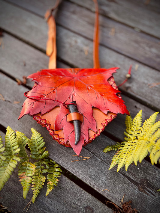 Ledertasche mit rot-orangefarbener Ahornblatt-Klappe und dunklem Knebelverschluss auf Holzplanken neben Farn.