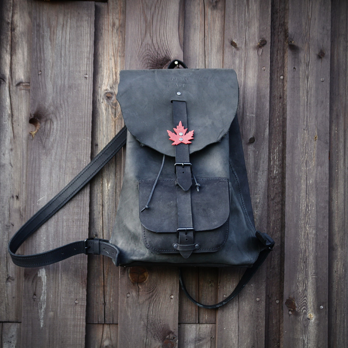 Schwarzer Lederrucksack mit rotem Ahornblatt, Fronttasche und verstellbaren Riemen vor einer Holzwand