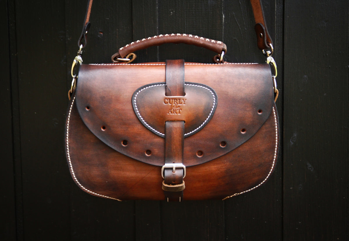 Vorderansicht der Leder-Umhängetasche Skadi mit Logo-Prägung, Lochdesign und Schnalle vor schwarzem Holz.