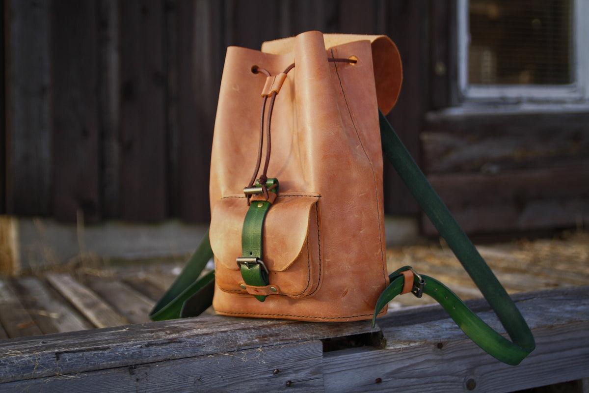 Heller Lederrucksack mit grünen Riemen, Fronttasche und hochgeklappter Verschlussklappe in seitlicher Ansicht