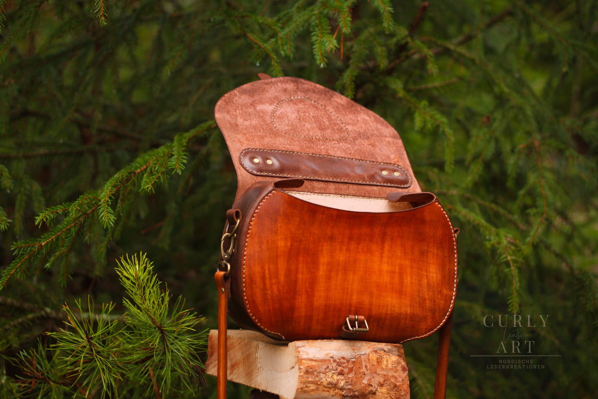Leder-Umhängetasche Skadi mit geöffneter Taschenklappe auf einem Holzstamm im Wald.