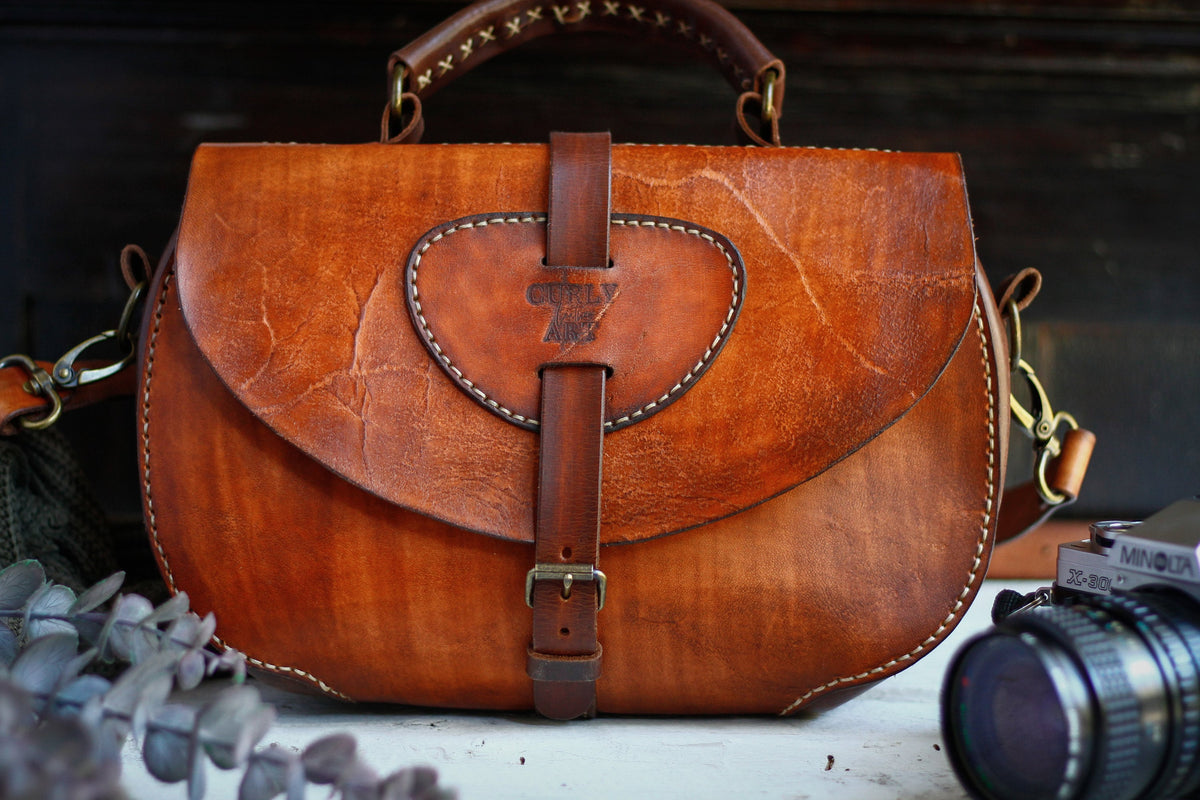 Vorderansicht der cognacfarbenen Ledertasche Skadi  mit rundem Patch, Schnallenverschluss und Tragegriff.