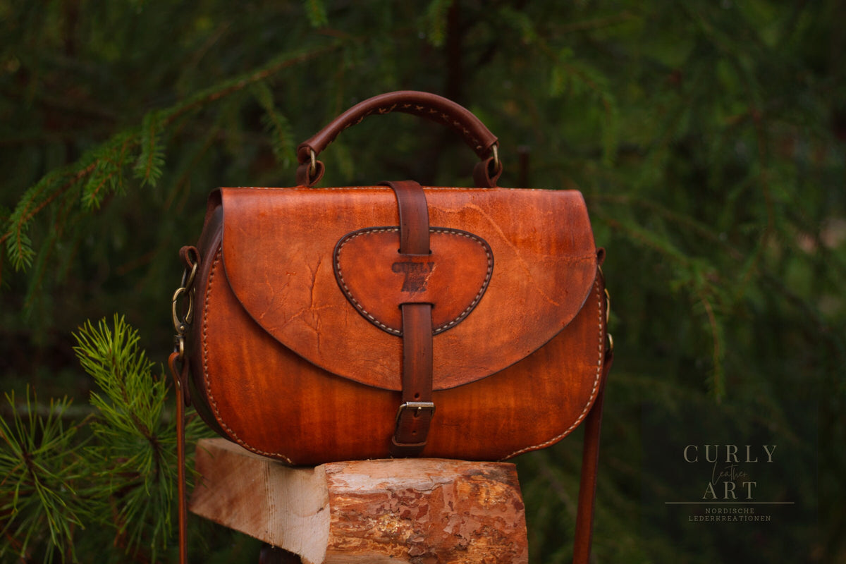 Ledertasche Skadi von vorne mit geschlossener Klappe und Schnallenverschluss auf einem Holzstamm.