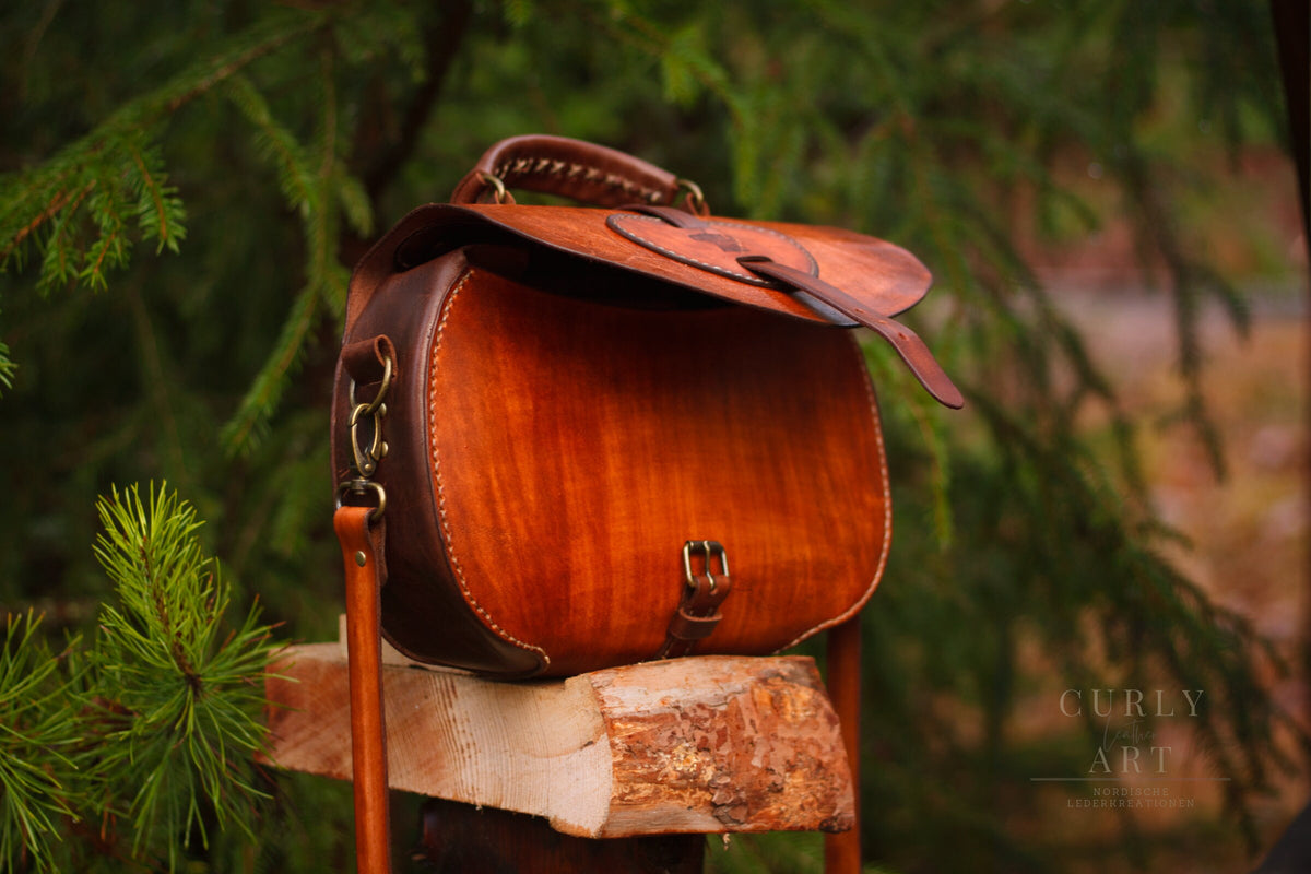 Schrägansicht der Ledertasche Skadi mit leicht angehobener Klappe und seitlicher Befestigung des Schulterriemens.